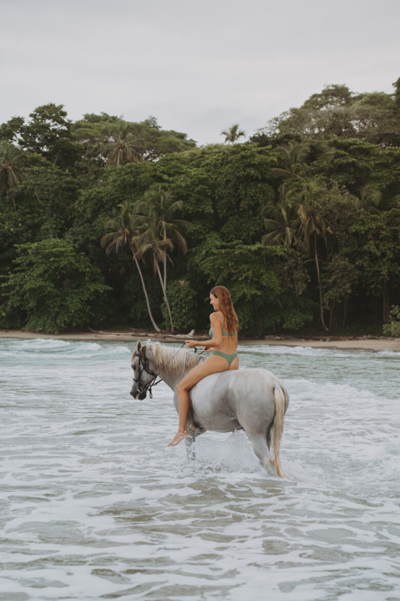 A woman in a green bikini rides a white horse through shallow ocean waves near a sandy beach lined with dense, tropical greenery. The sky is overcast, and the tranquil scene captures the peaceful interaction between the rider, her horse, and nature.