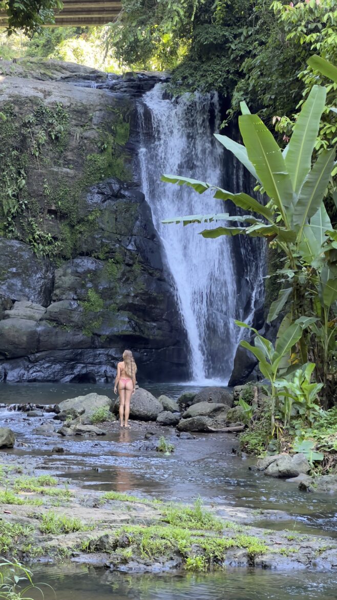 A women with long hair stands on rocks in front of a waterfall surrounded by lush greenery. Large leaves of various plants, including banana plants, are visible in the foreground and background. The scene depicts a tranquil and natural environment.