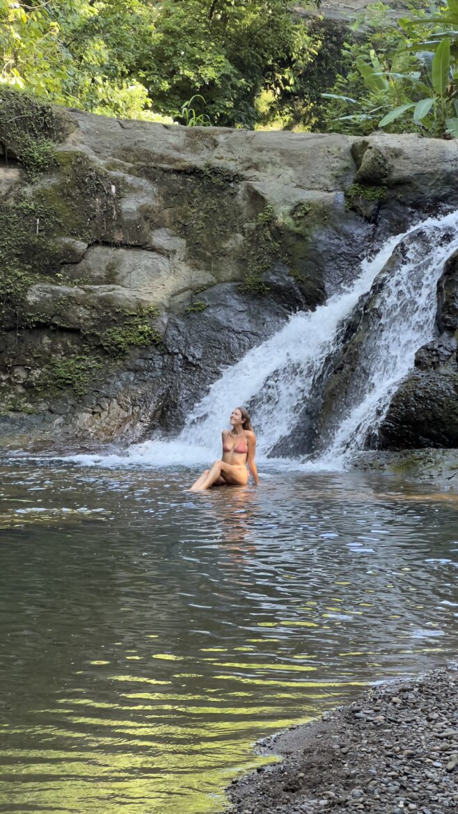 A person in a swimsuit sits on a rock beneath a small, cascading waterfall in a lush, green environment. The water flows gently around them, creating a serene and peaceful scene. The background is filled with rocks and greenery.